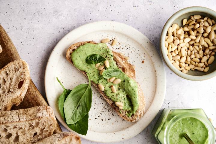 A slice of whole grain bread topped with green basil pesto, pine nuts, and fresh basil leaves, beside a bowl of pine nuts.