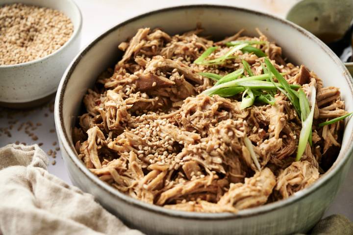 Shredded slow-cooked sesame ginger chicken topped with sesame seeds and sliced green onions in a ceramic bowl, with a bowl of sesame seeds in the background.