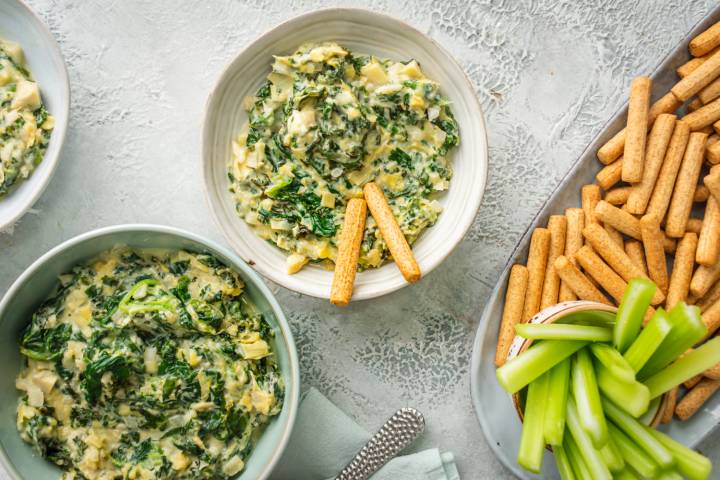 Bowls of creamy spinach, artichoke, and kale dip served with crispy breadsticks and fresh celery sticks for dipping.