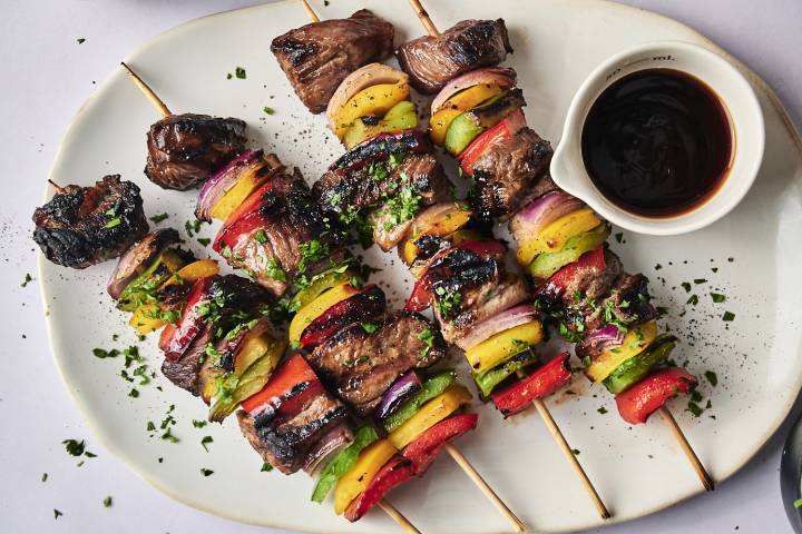 Grilled kebabs featuring marinated beef and colorful vegetables, served with a small bowl of dipping sauce on a white plate.