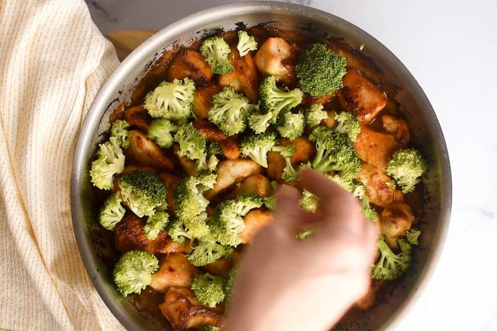 Broccoli being added to a skillet with orange chicken.