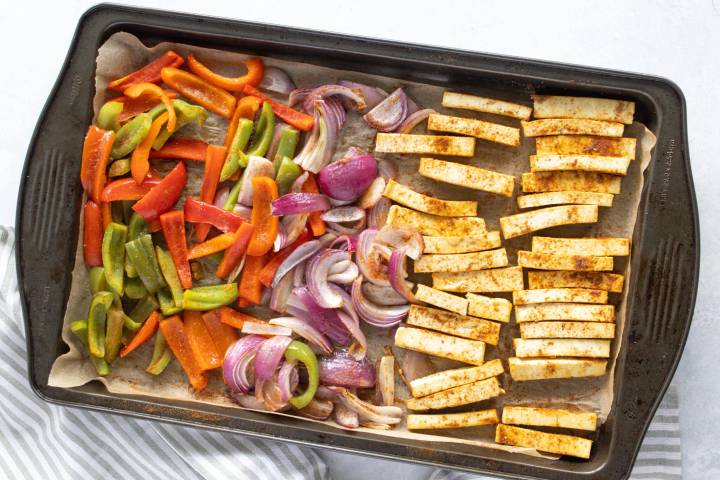 Cooked tofu and fajita seasoning on a sheet pan with sliced bell peppers and red onions.