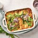 A close-up of cheesy chicken enchiladas being served from a casserole dish, topped with avocado, red onion, jalapeños, and fresh cilantro.