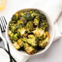 Bowl of roasted broccoli topped with grated cheese, set on white napkin. Two black forks lie beside, with a glass of yellow liquid nearby.