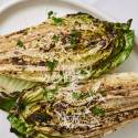 Grilled romaine lettuce halves topped with grated Parmesan, herbs, and black pepper, served with a lemon slice on a white plate.