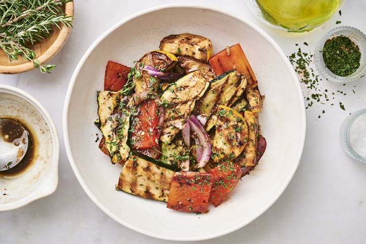 A colorful bowl of grilled vegetables, including zucchini, bell peppers, and onions, garnished with herbs and drizzled in dressing.
