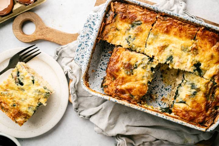 Golden-brown spinach and cheese casserole in a speckled baking dish, partially cut. A single slice on a white plate with a fork, set on a wooden board.
