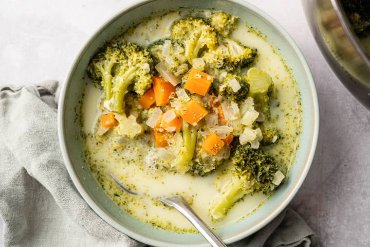 A bowl of broccoli cheddar soup with visible broccoli florets, diced carrots, and onion chunks in a creamy broth. A spoon rests on the bowl's edge. The soup sits on a light-colored cloth, conveying a comforting, homemade feel.