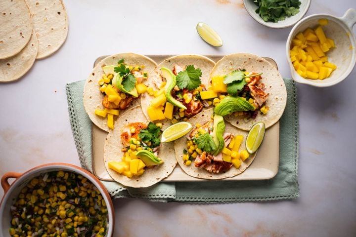 Salmon tacos with corn salsa, mango, fresh avocado slices, and lime wedges on a tray. 