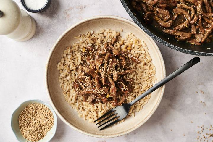 Bowl of sesame beef served over brown rice and sprinkled with sesame seeds.