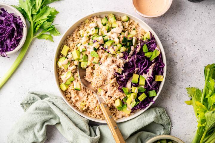 A colorful bowl of brown rice topped with cubed avocado, diced cucumber, purple cabbage, and dressing. Celery and a linen cloth are nearby, creating a fresh and vibrant look.