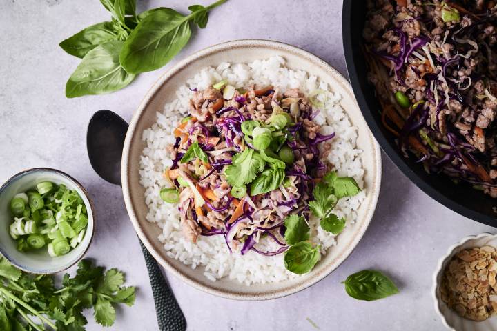 A bowl of white rice topped with sautéed ground meat, purple cabbage, and green herbs, beside a bowl of chopped scallions and fresh basil.
