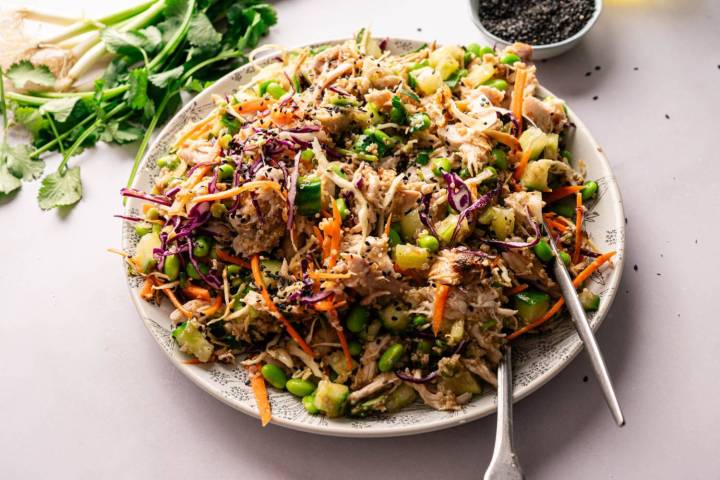 A vibrant Asian salad with shredded chicken, colorful cabbage, edamame, carrots, and cucumbers. Garnished with black sesame seeds. Fresh cilantro and green onions are nearby.