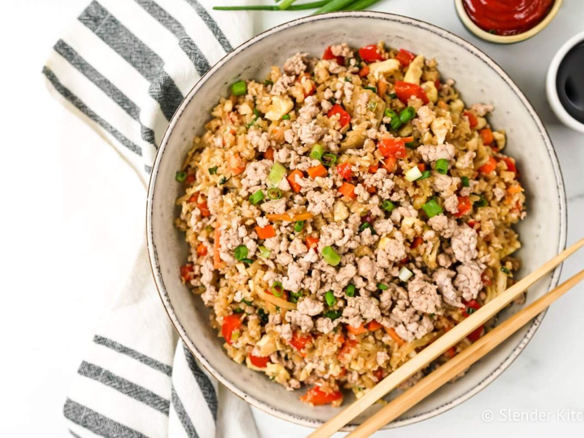 Cauliflower rice with ground pork, cabbage, carrots, and green onion in a bowl with chopsticks.